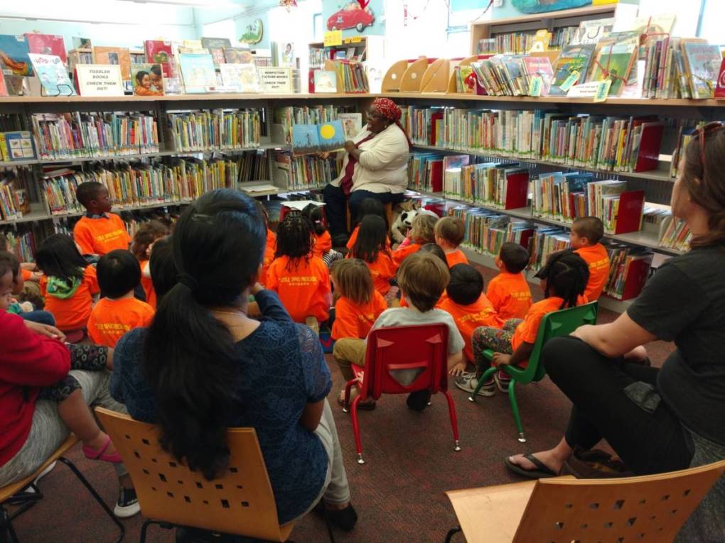 A woman in a white sweater sits in front of a group of children reading a book.