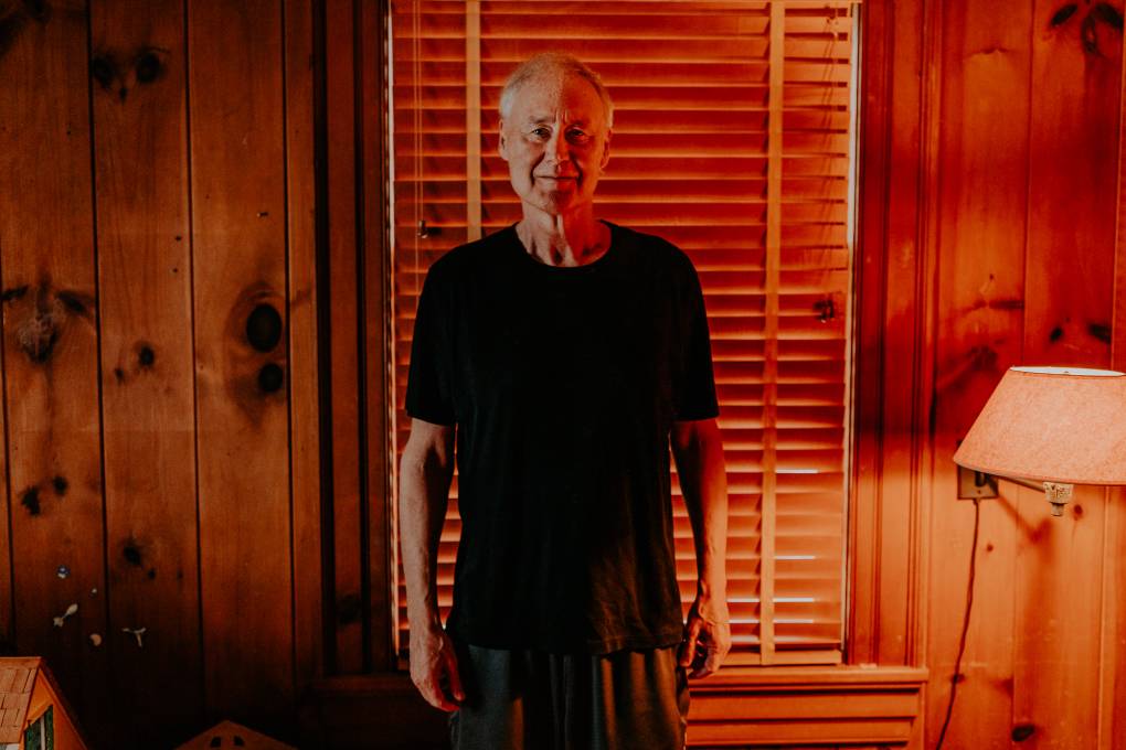 An elder white man in a black t-shirt stands in front of a window with closed blinds.
