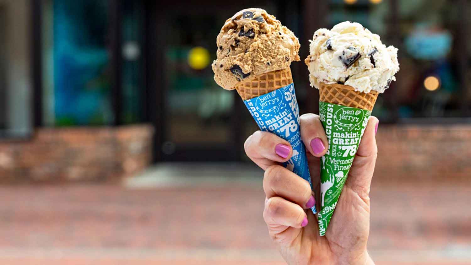 A woman's hand with pink painted nails holds two cones of ice cream in the air