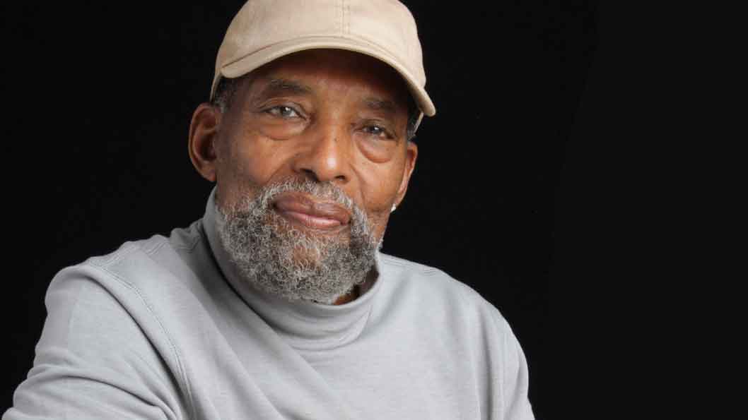 A smiling Black man in a ballcap and greying beard, wearing a grey sweatshirt.