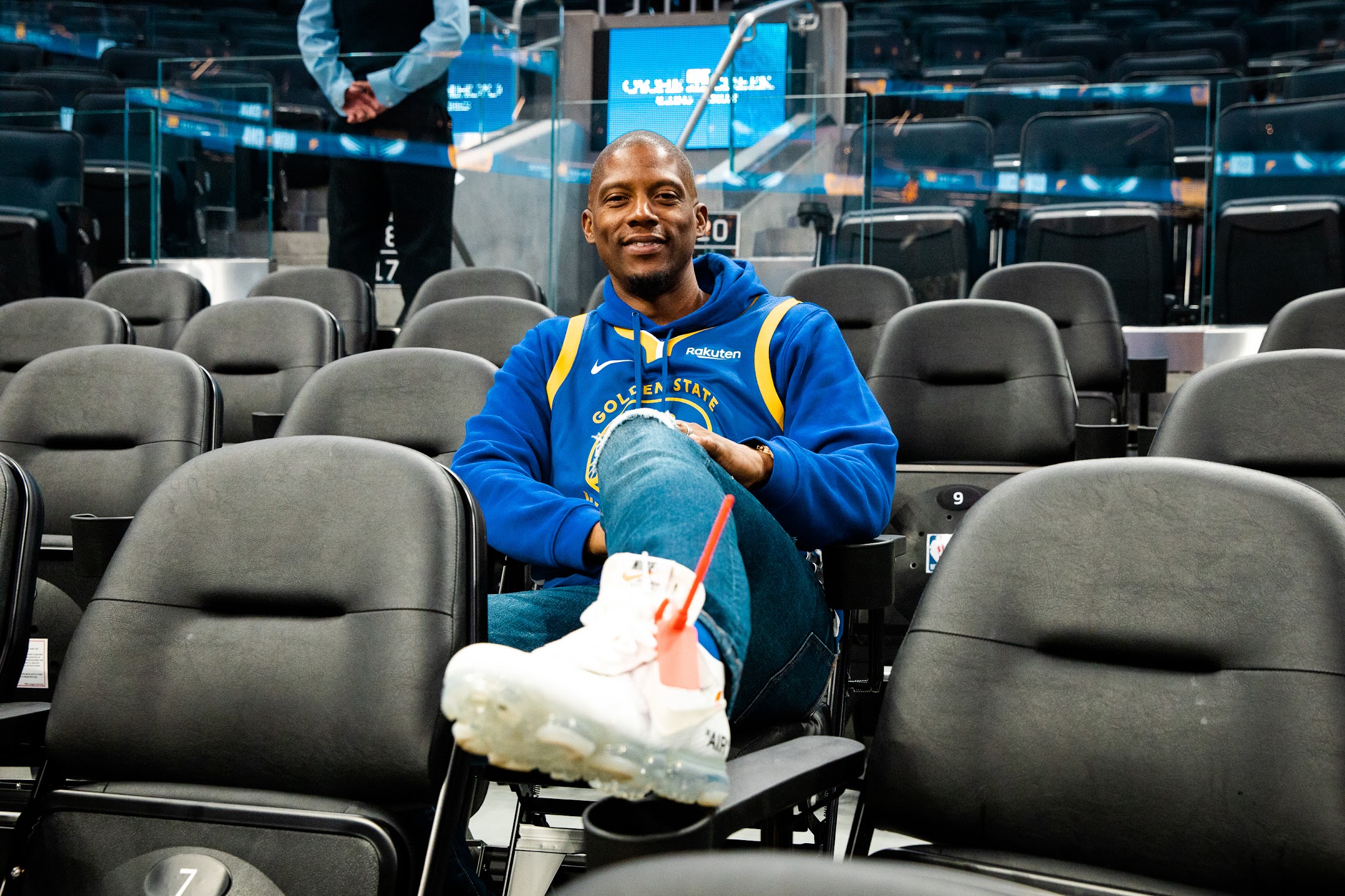 A man sits in a row of black leather chairs, with one foot up and smiling.