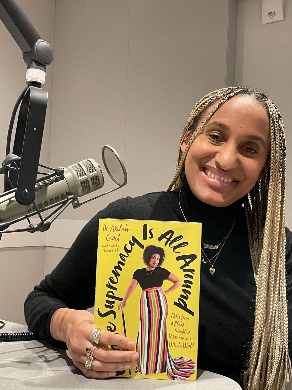 Dr. Akilah Cadet poses while sitting at KQED studios and holding a copy of her book, 'White Supremacy Is All Around: Notes from a Black Disabled Woman in a White World'.