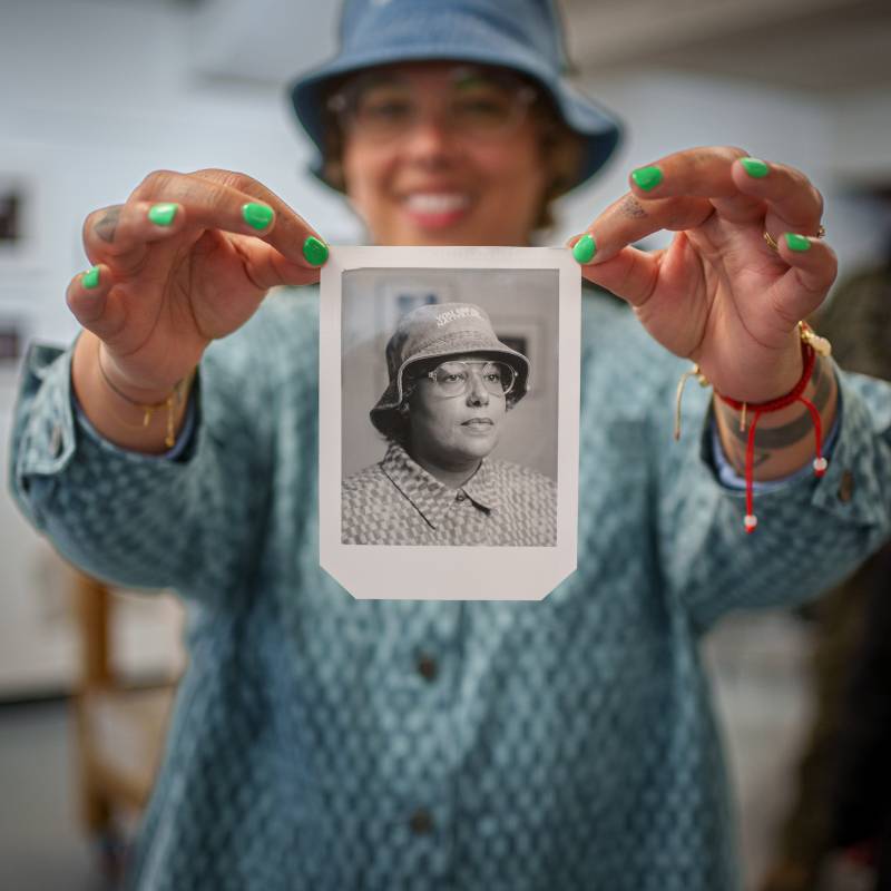 A person in a bucket hat and dress shirt hold up a printed photo of themselves. 