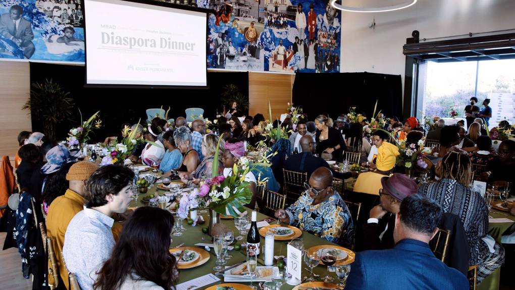The crowded dining room at an elegant gala. The text projected onto a screen reads, "Diaspora Dinner."