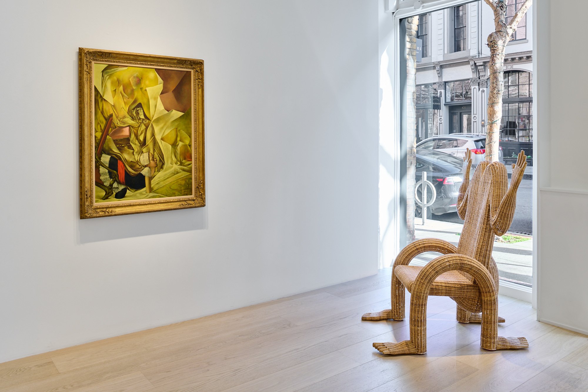 Framed abstract painting on wall, wicker hair with human-like arms and legs in front of it