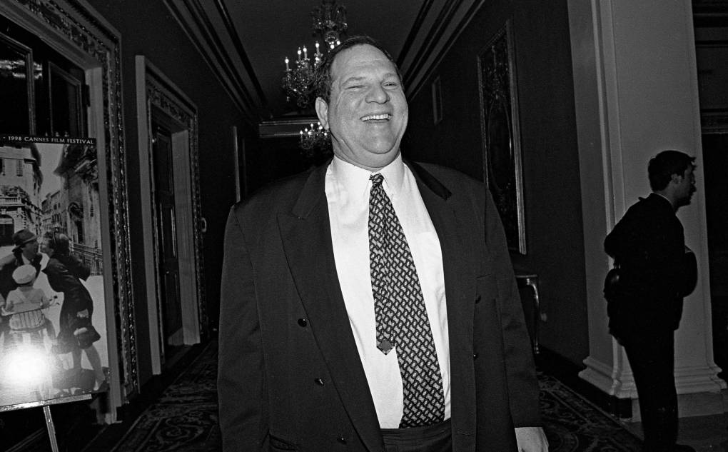A large white man in a suit stands in a grand hallway laughing.