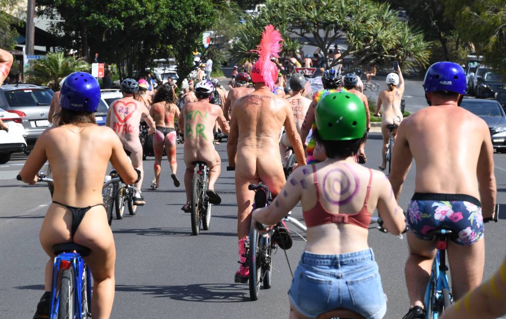 A group of men and women in varying stages of nudity ride bikes together up a road.