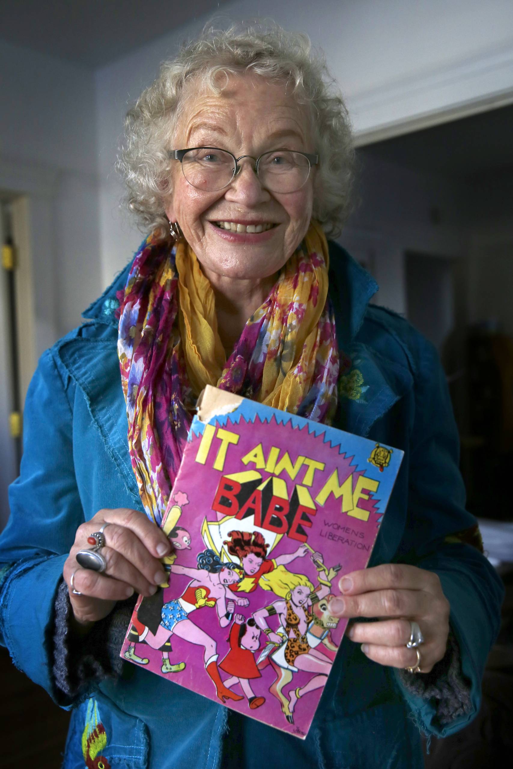 A senior white woman smiling as she holds up a comic book titled 'It Aint Me Babe.’