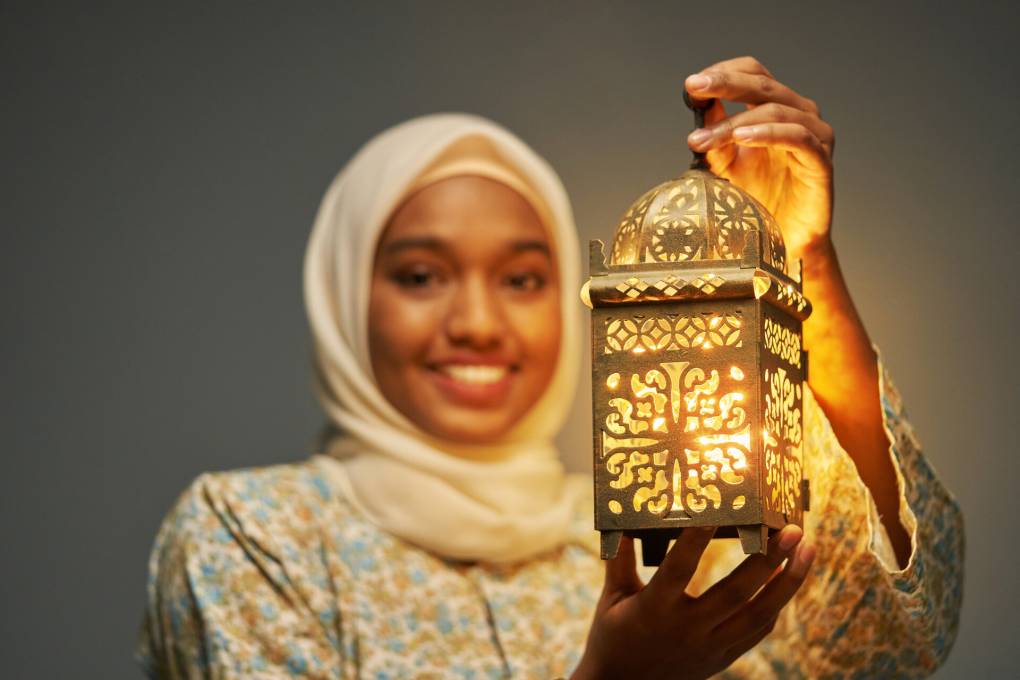 A young woman wearing a light-colored hijab smiles and holds a lit lantern up to the camera