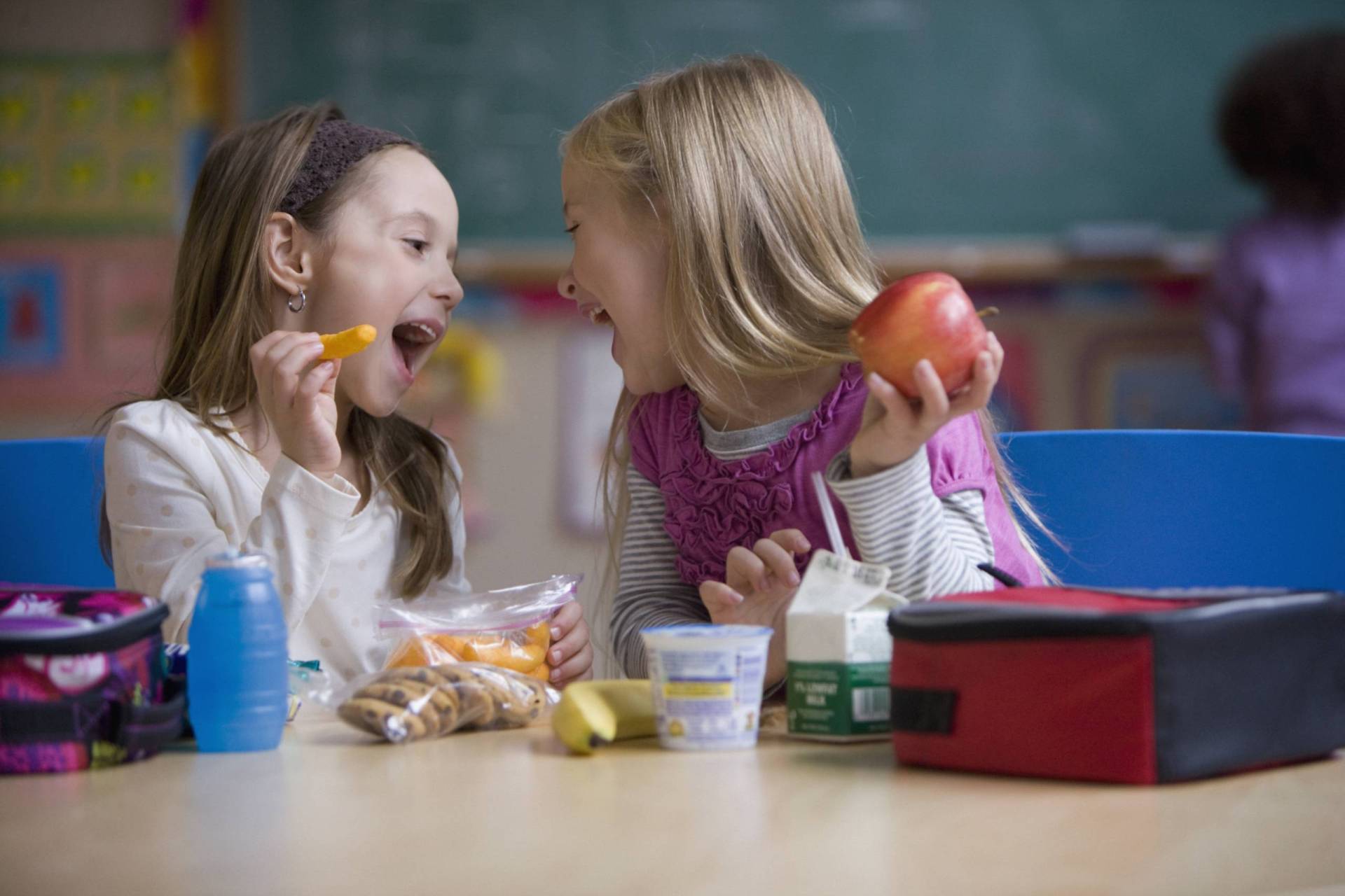 Two elementary school girls sit with lunch boxes in a classroom, pulling happy faces at one another.