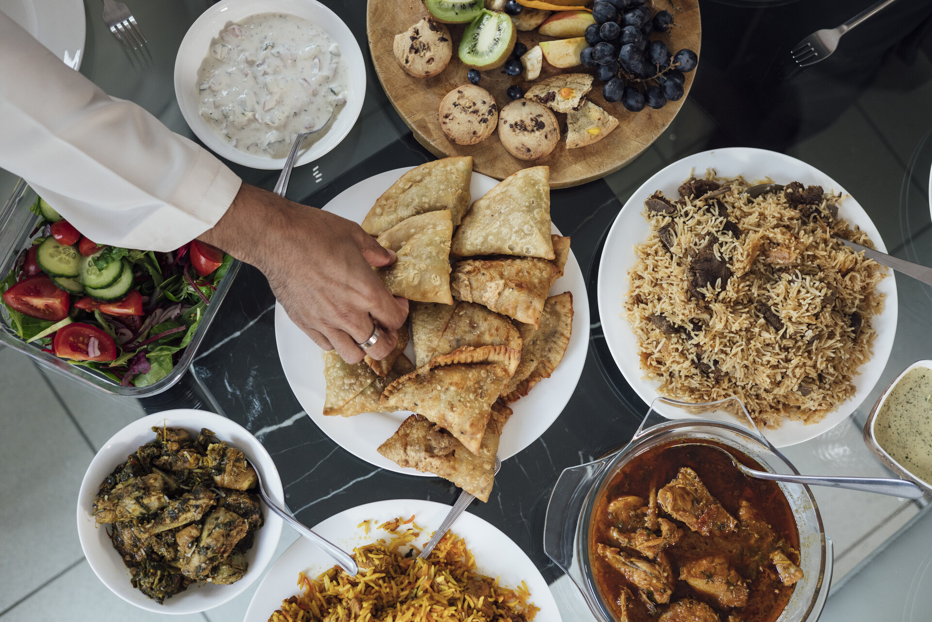 An overhead shot of a delicious-looking array of plated foods on a gray table, including samosas and curry, with a hand reaching in to take some