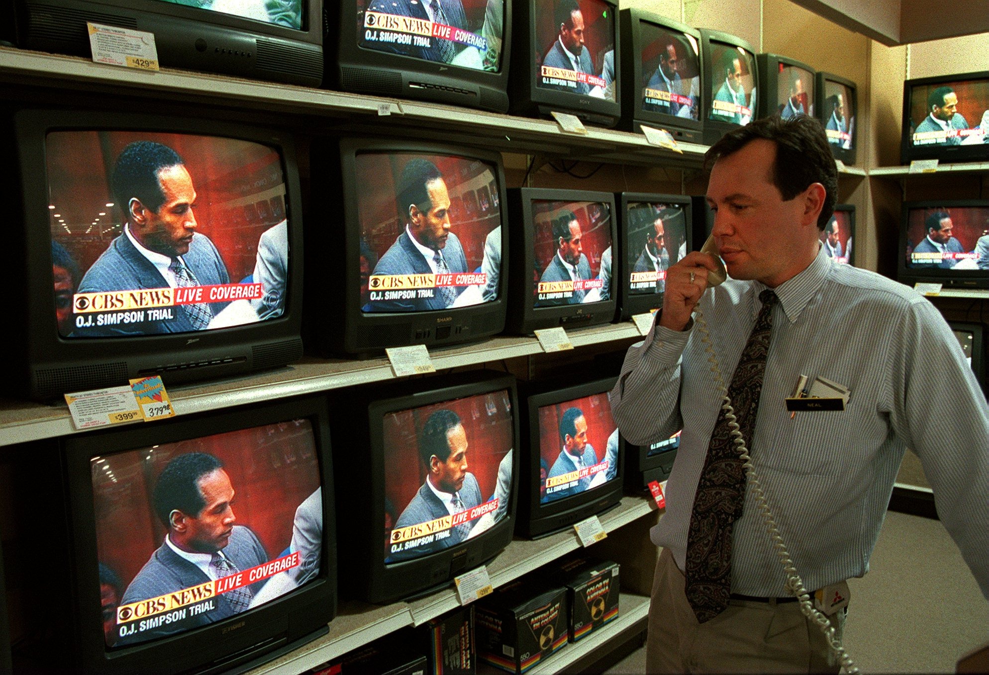 A man in a shirt and tie talks on a 1990s-era phone while standing in front of a wall of televisions all showing the trial of OJ Simpson.