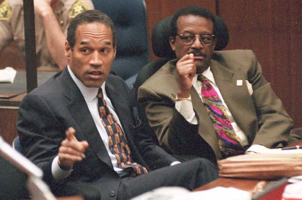 OJ Simpson sits in court, gesturing to someone off camera. At his side sits one of his attorneys.