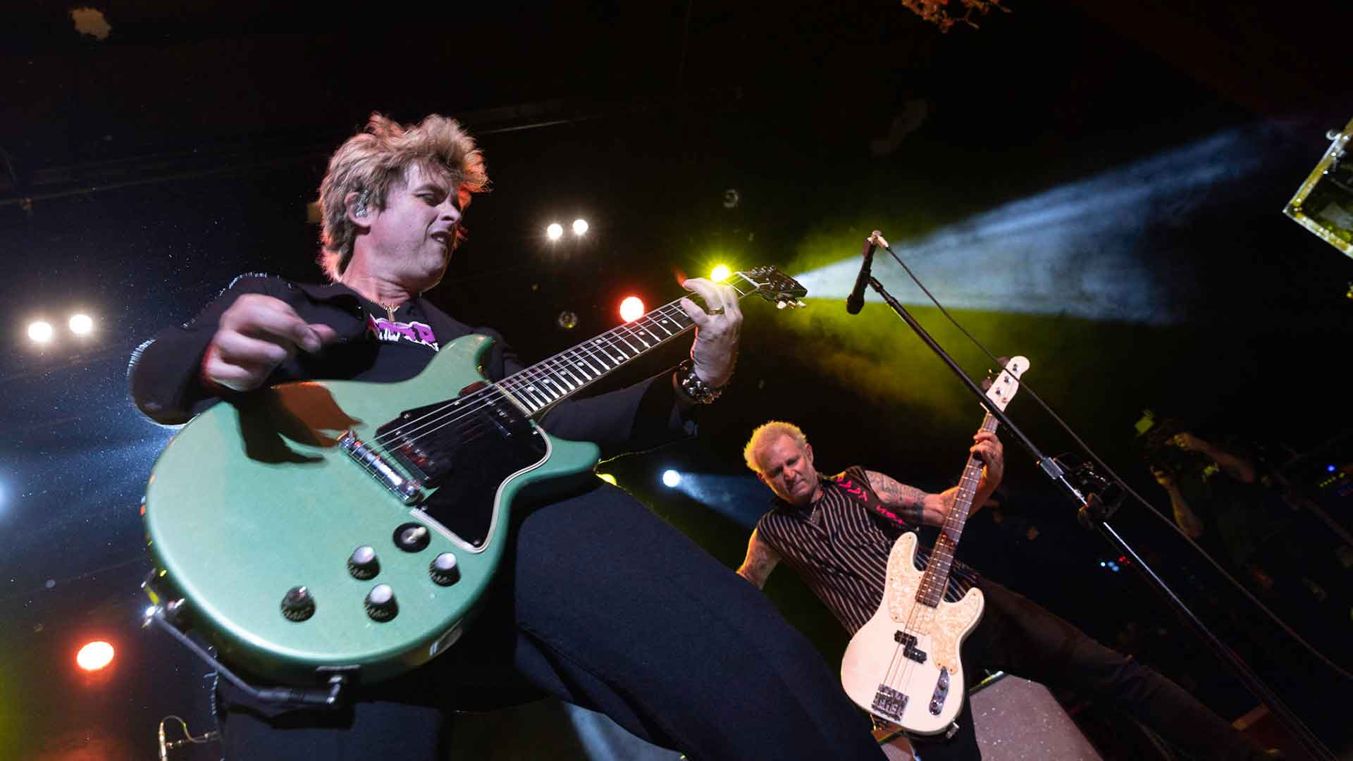 A man plays a Les Paul-style guitar, photographed from below, with a man playing bass and stage lights in the background