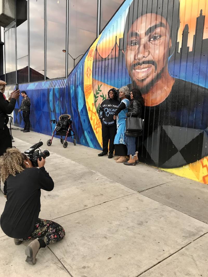 Woman with video camera kneels down to document three individuals standing in front of a mural.