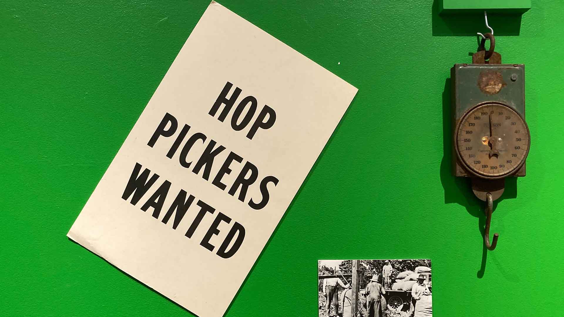 A rusty scale sits against a green background, next to a sign that reads "Hop Pickers Wanted"