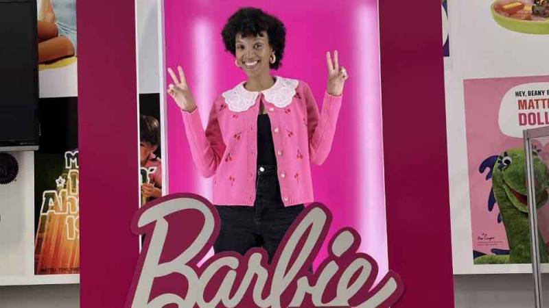 A Black woman in a pink sweater flashes two peace signs while inside a life-sized Barbie box