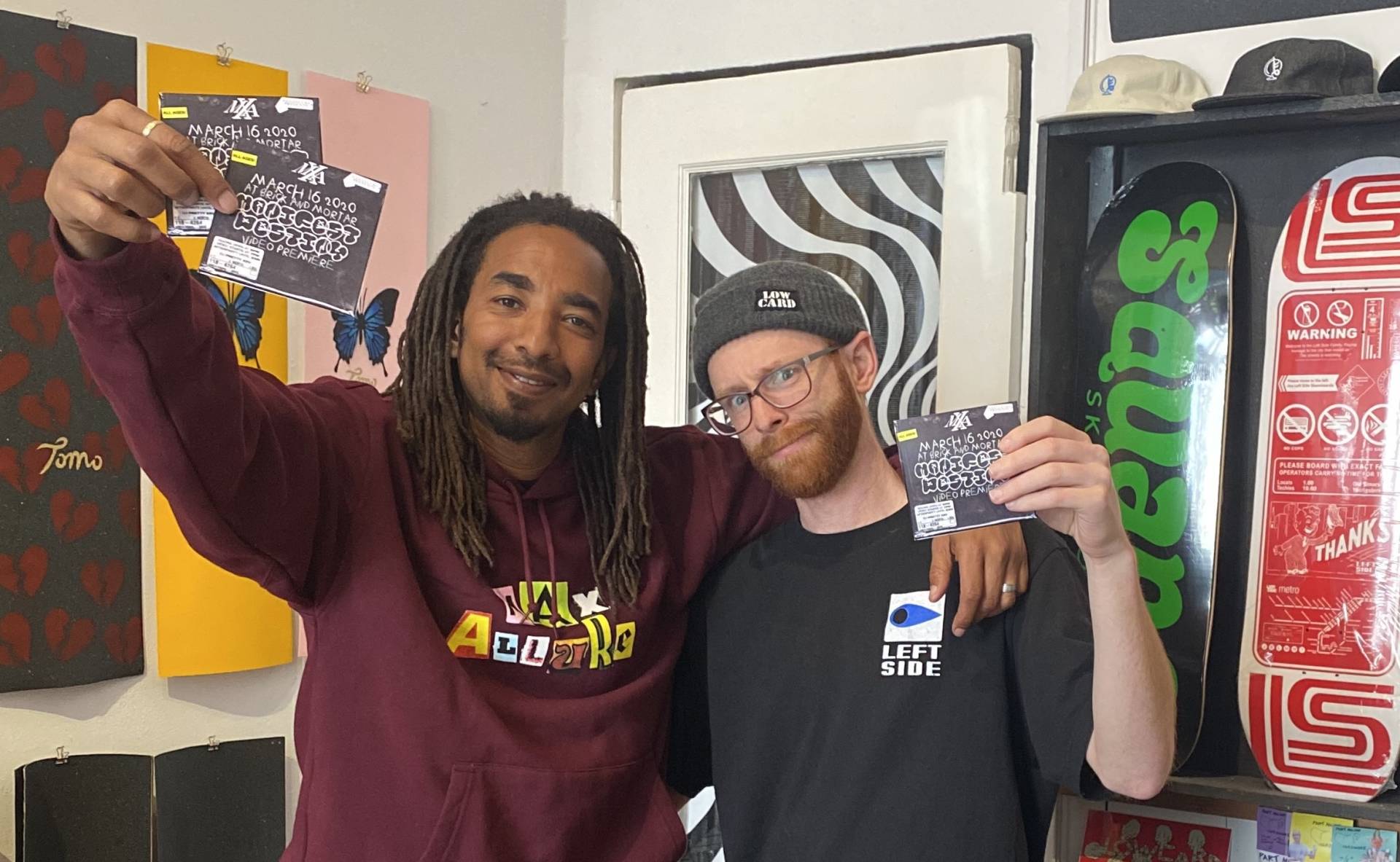 A Black man with long locs stands with his arm around a white man with beard and glasses inside a skateboard shop. They are both smiling.