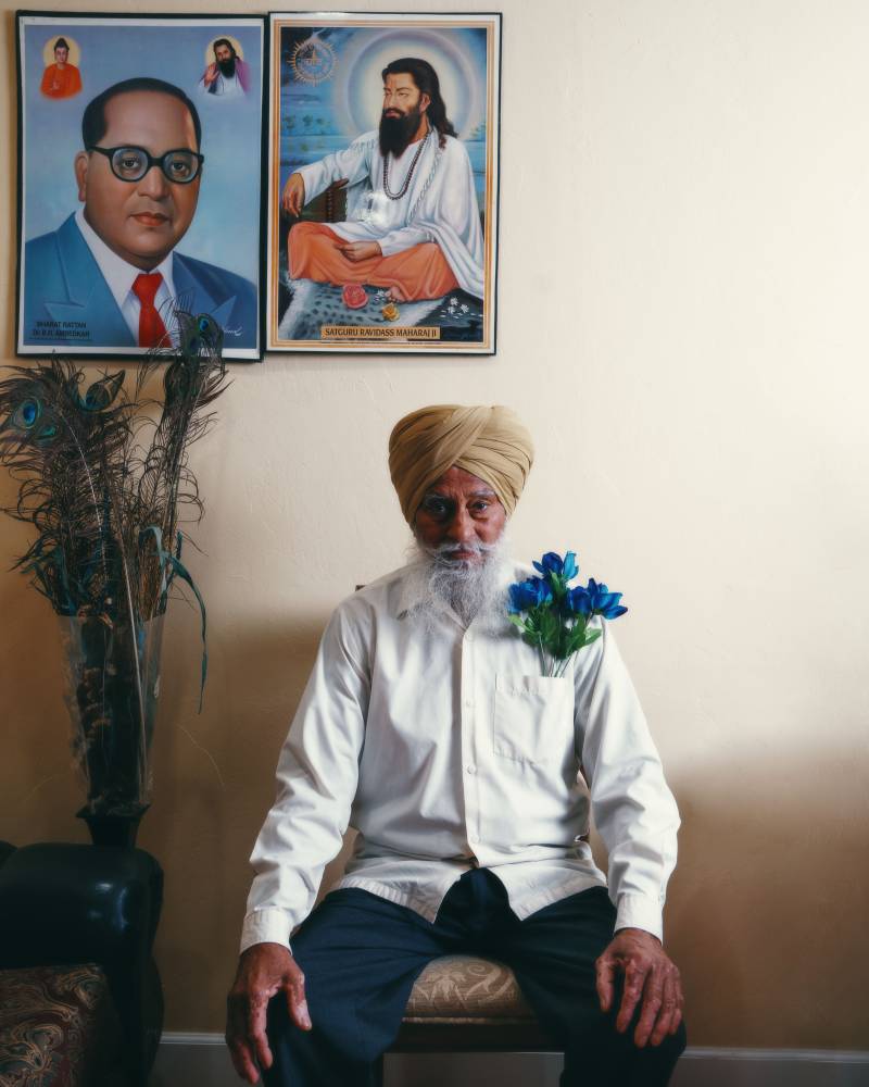 An elder man wearing a turban sits in front of a wall with two paintings on it.
