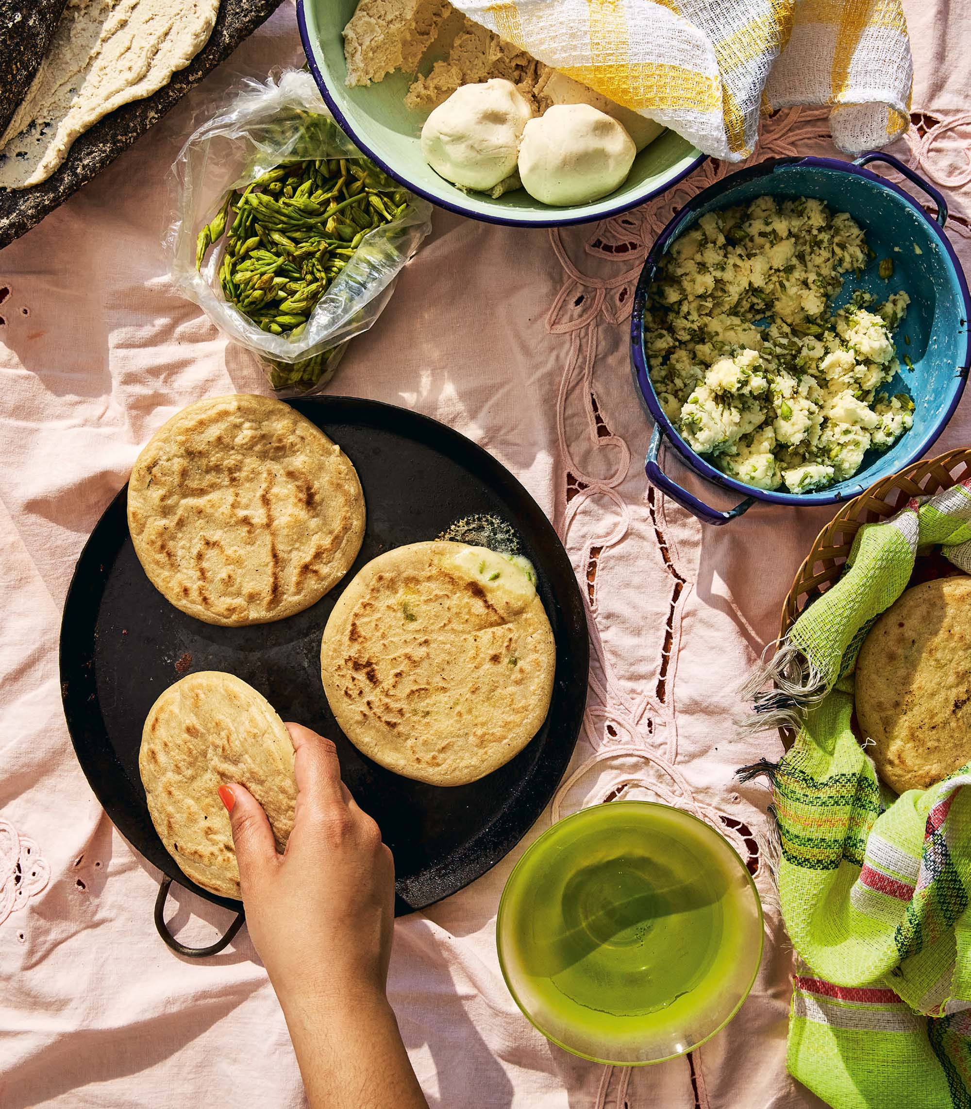 A hand picks up a pupusa off a black skillet. On the table, there's a bowl of cheese and loroco — the fillings for the pupusas.