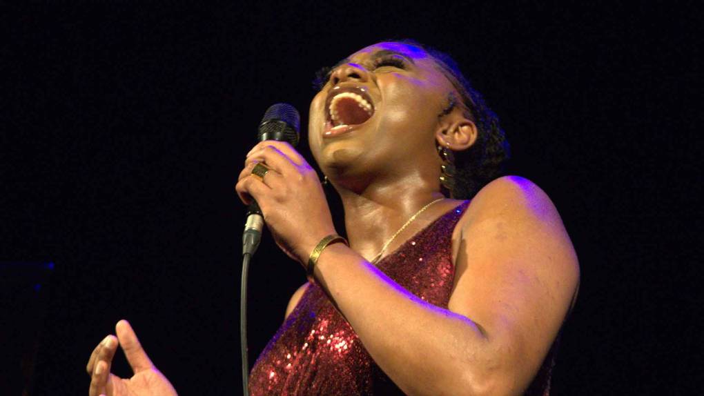 A black woman in a red dress sings into a microphone while tilting her head upward, eyes closed
