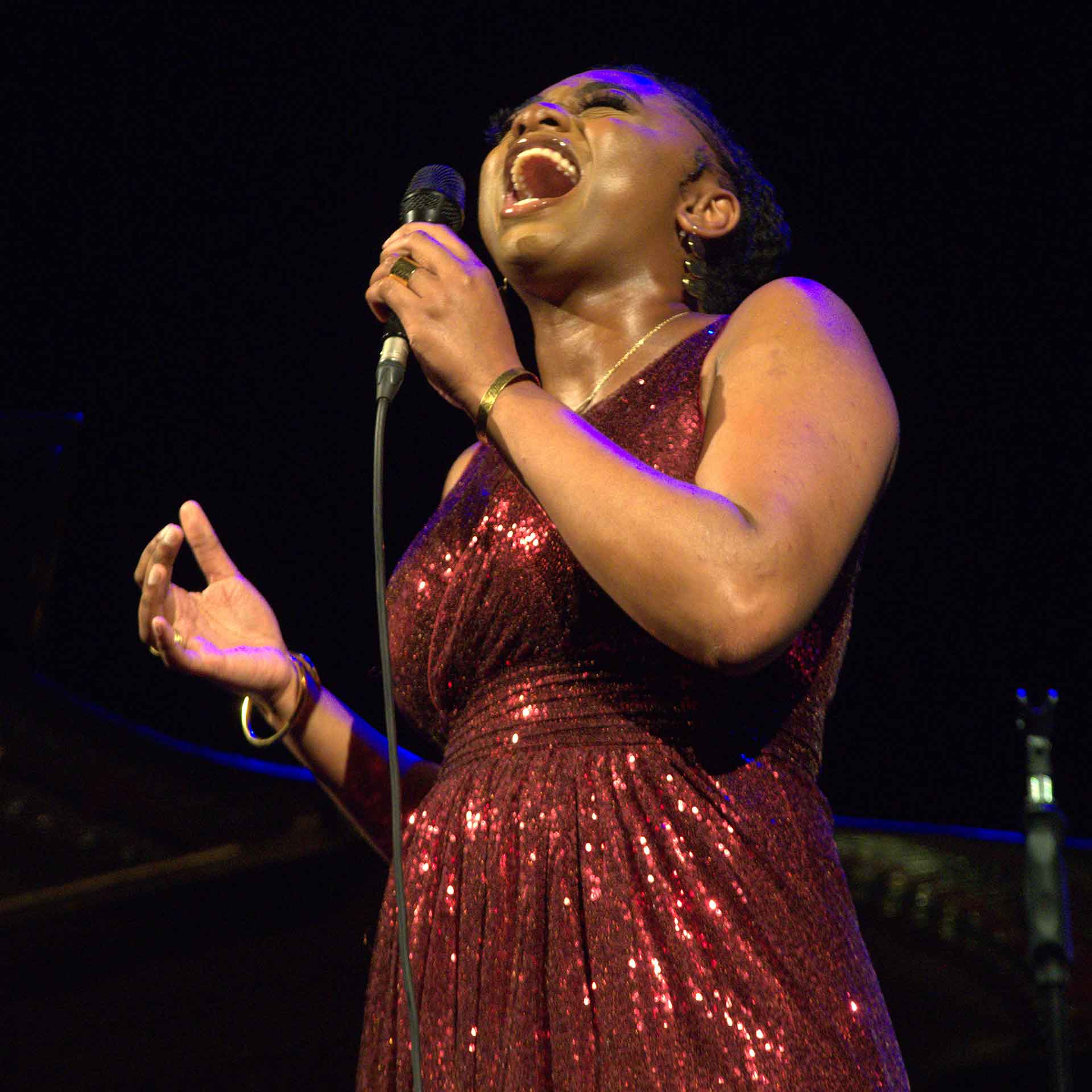 A black woman in a red dress sings into a microphone while tilting her head upward, eyes closed