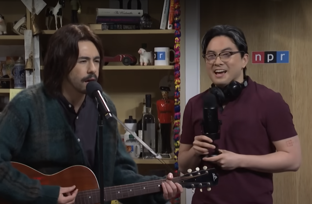 A man with long brown hair plays an acoustic guitar and sings into a microphone while a man stands nearby holding a radio mic and talking, headphones around his neck. They are in an office setting.