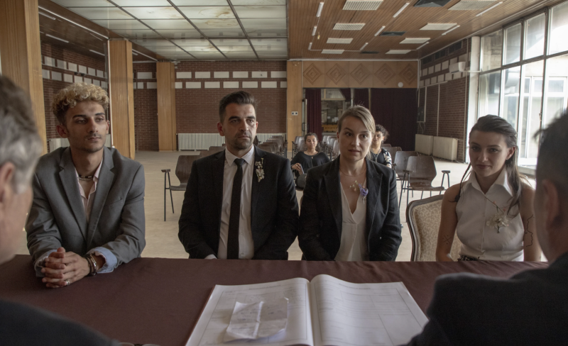 A man and woman sit wearing formal clothes in front of a desk in a courtroom. A younger man and woman sit either side of them.