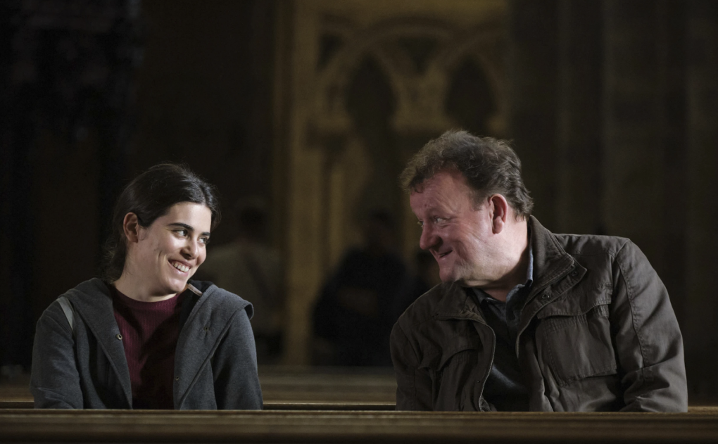 A young Syrian woman smiles at an older white man. They are sitting on a church pew.