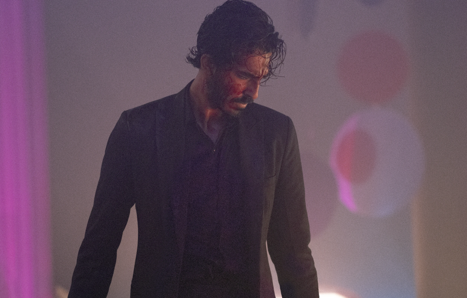 A South Asian man in a suit stands in dim light, his face smeared with blood.
