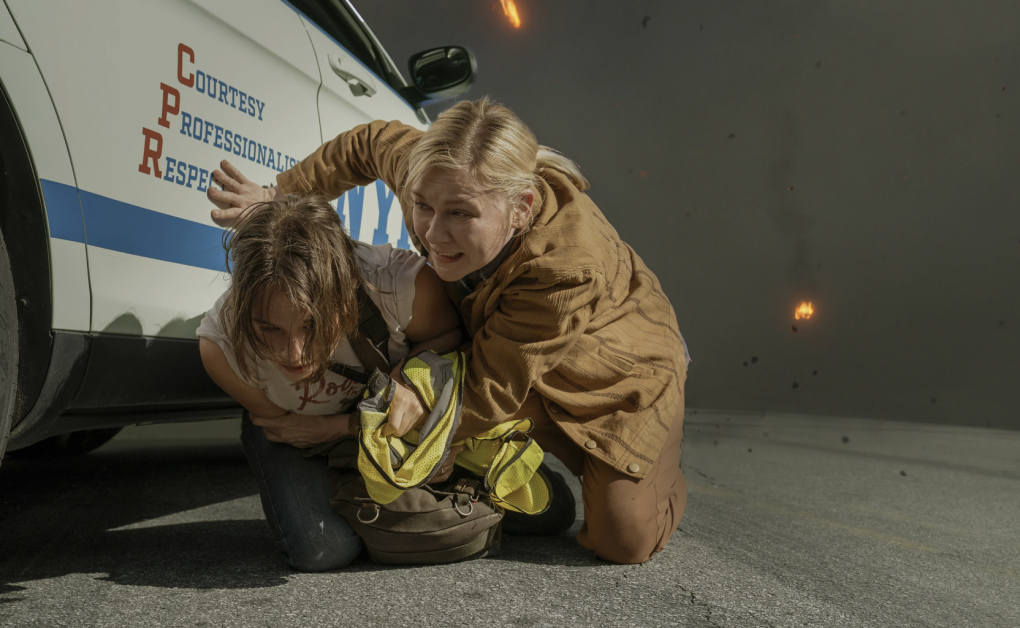 Two white women crouch close to the side of a police car while fire falls in the smoky distance.