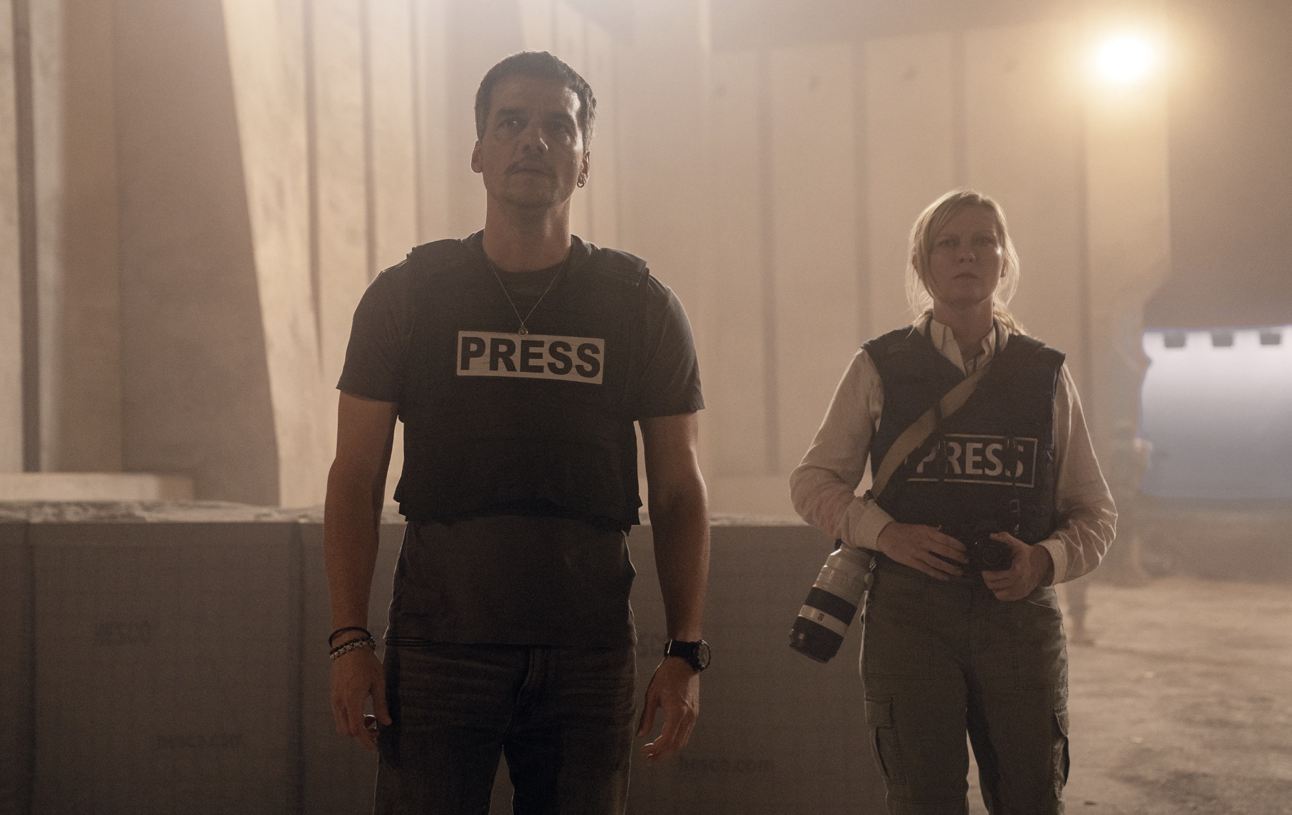 A man and a woman wearing a large camera stand in a smoky room, both wearing vests marked PRESS.