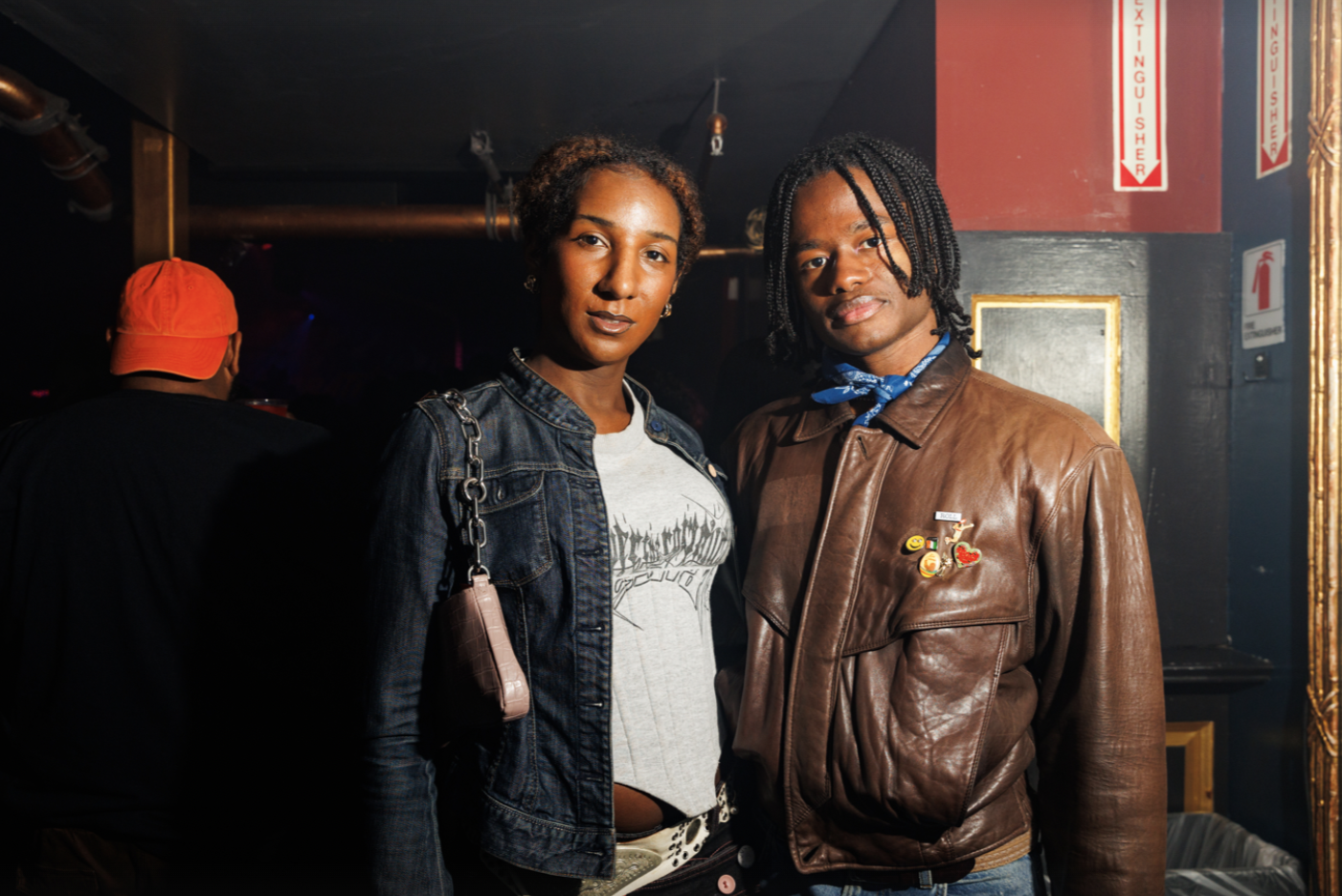 Two young people of color stand side by side inside a nightclub with thoughtful expressions on their faces.