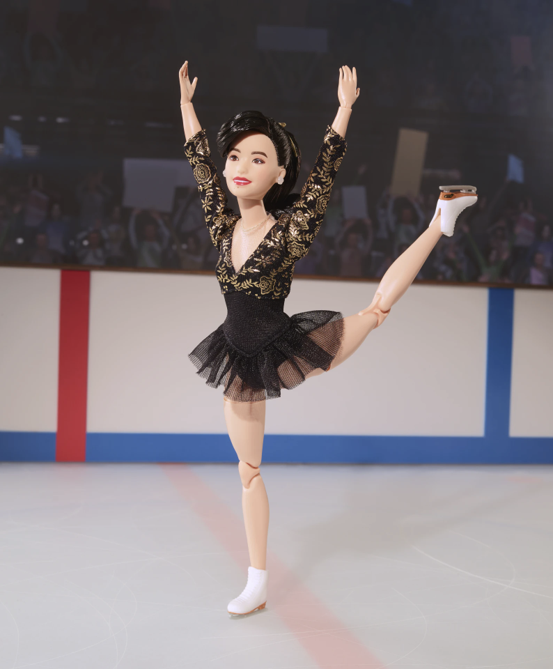 A doll of an ice skater stands, one leg stretched out and arms raised on the ice. The doll is wearing a black lace and chiffon costume. 