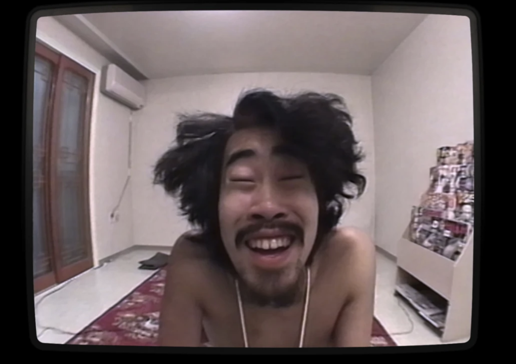An unkempt, shirtless Japanese man in a sparse white room leans towards the camera and smiles.