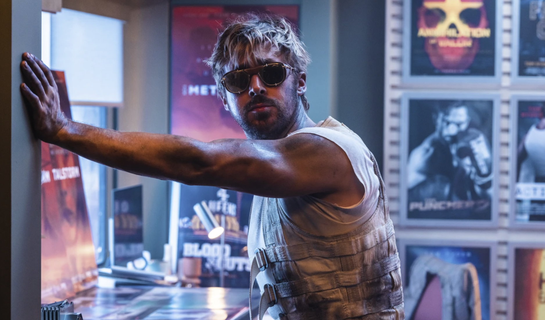 A handsome but disheveled man wearing sunglasses leans one muscular arm against a wall. Movie posters line the wall behind him.