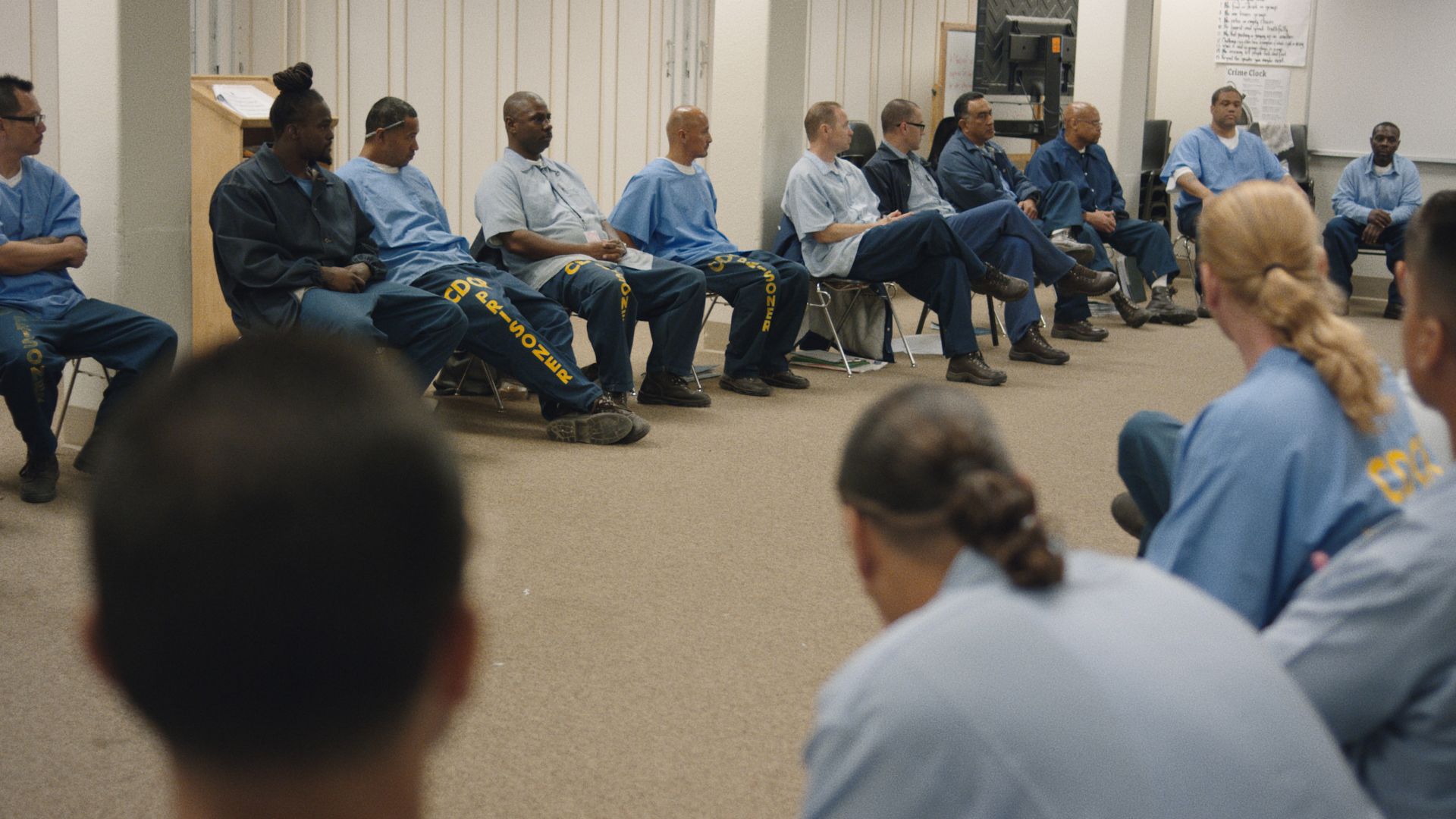 A scene from the film, 'The 50,' shows a room where men are dressed in state-issued prison clothing while sitting in a circle of chairs and holding a discussion. 