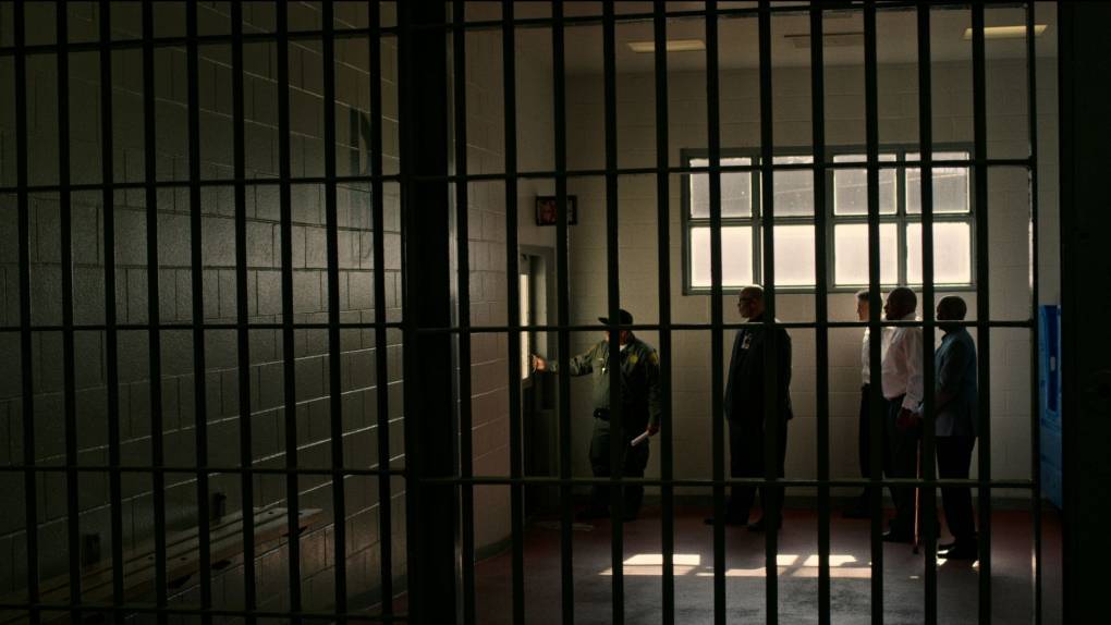 Prison bars in the foreground and shadows of men walking through a dark hallway in the background.