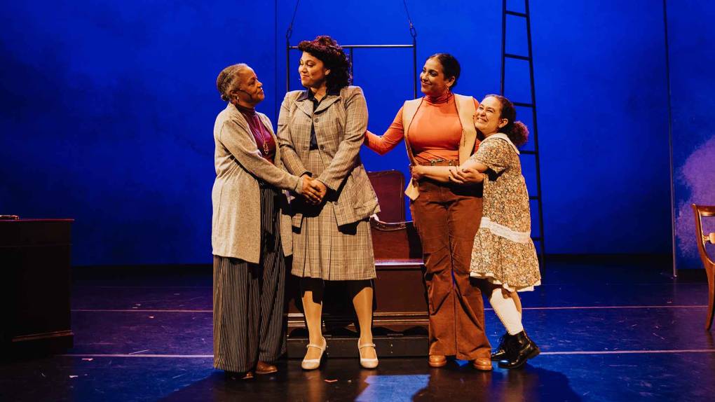 Four women stand on a stage backlit by blue, looking admiringly at each other