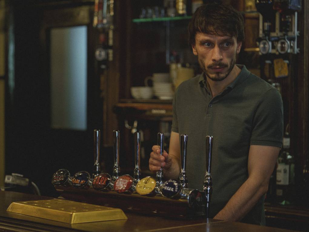 A sheepish looking white man stands behind the bar of a pub.