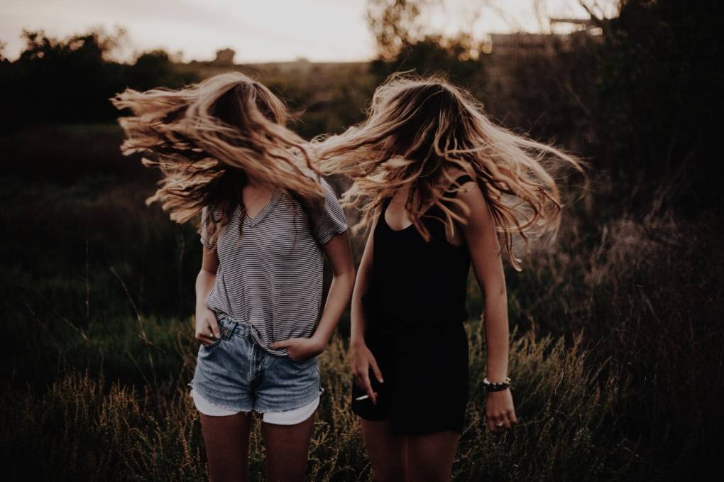 Two slender blonde young women stand in a field shaking their long hair in front of their faces.