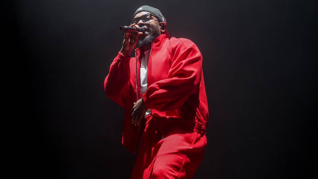 A bearded Black man in a red track suit and white t-shirt performs on stage.