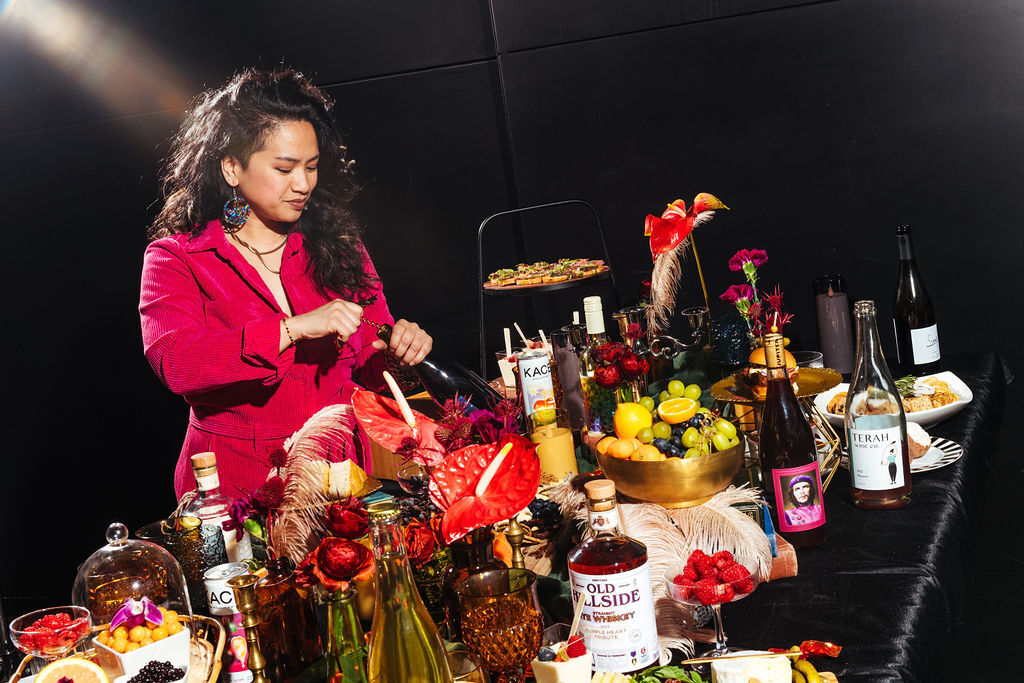 A woman in a red jacket stands before a lavish spread of drinks, appetizers and flowers.