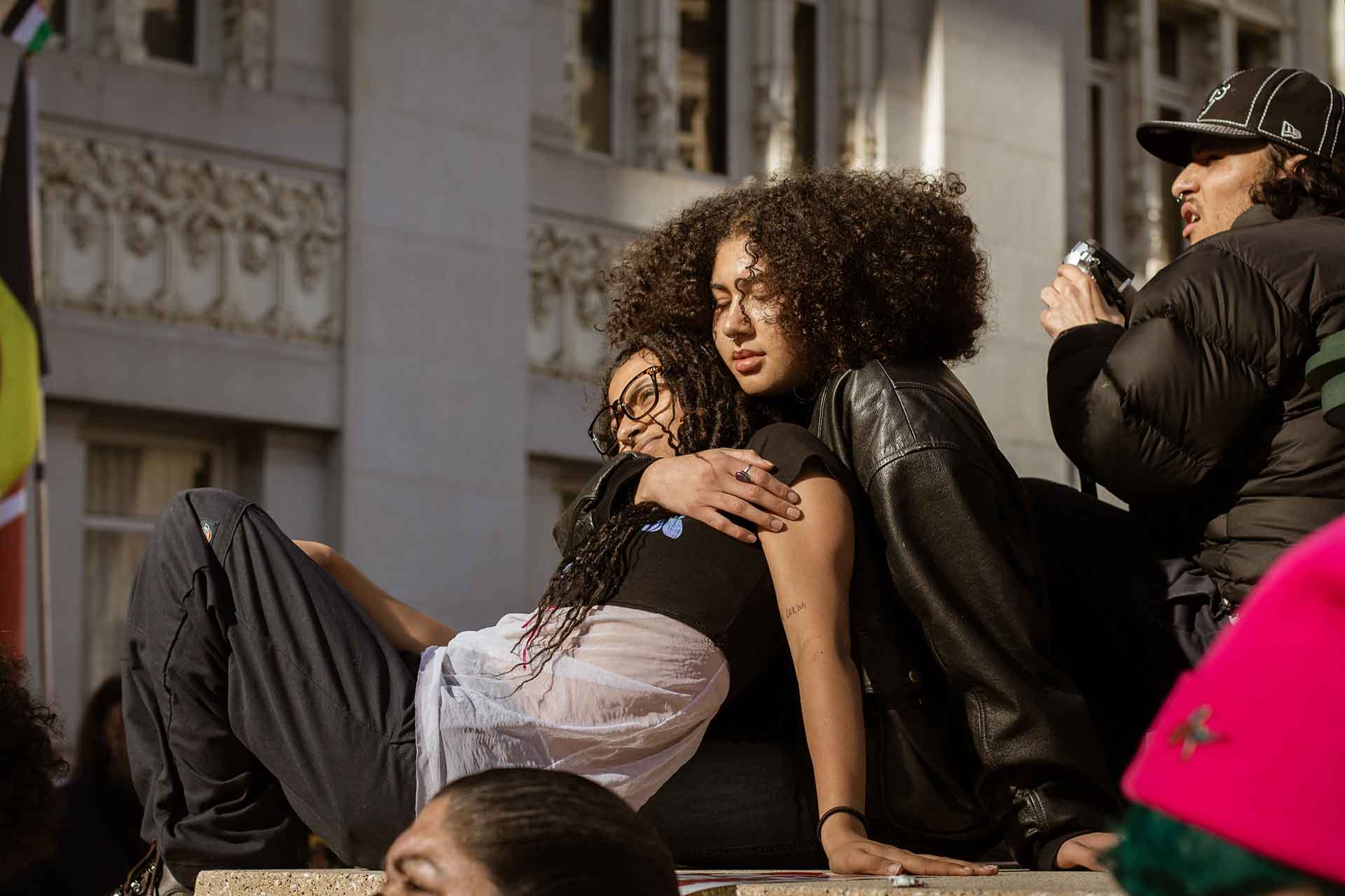 A young woman embraces another with a tall building in the background