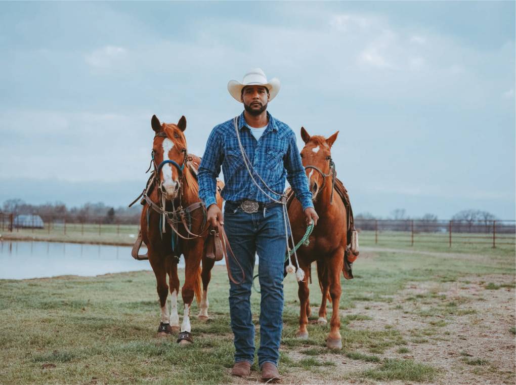 A Black cowboy leads two brown horses across an empty field.