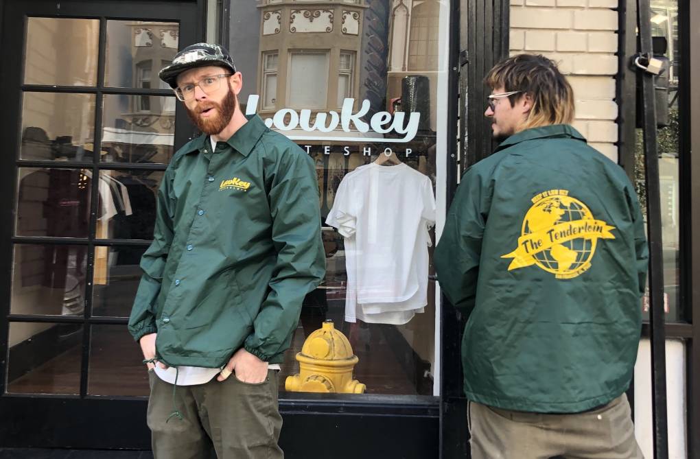 A white man with a red beard stands outside a shop front wearing a green windbreaker. Another man, with long half-bleached hair, stands to his right with his back turned. He is wearing the same windbreaker. It says 'The Tenderloin.'