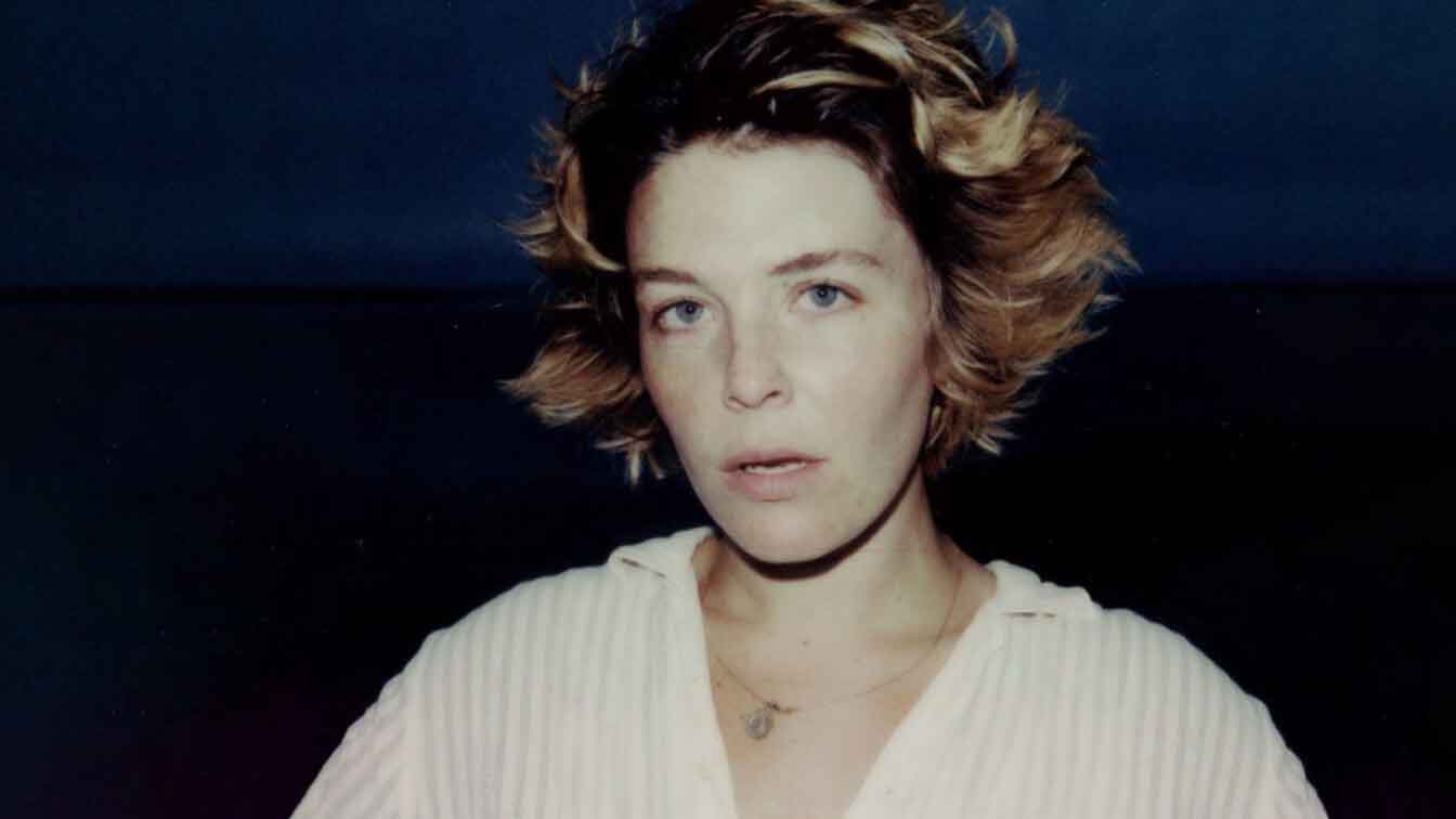 A woman in tousled hair and a white top looks at the camera, mouth slightly open
