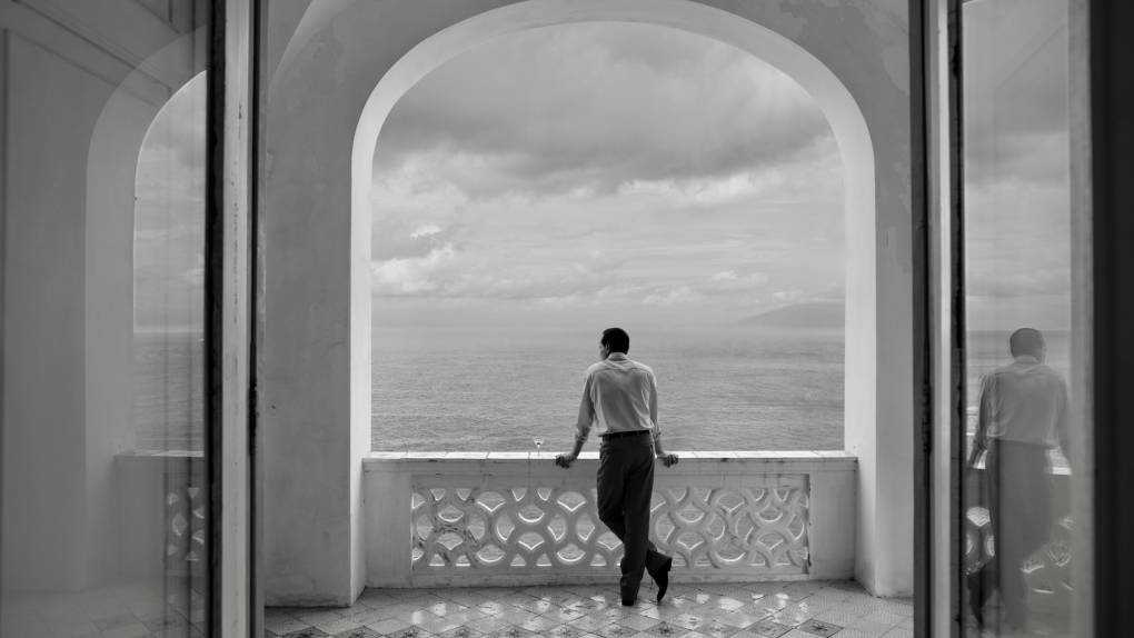 A man, viewed from behind, stands on an ornate veranda admiring the view.