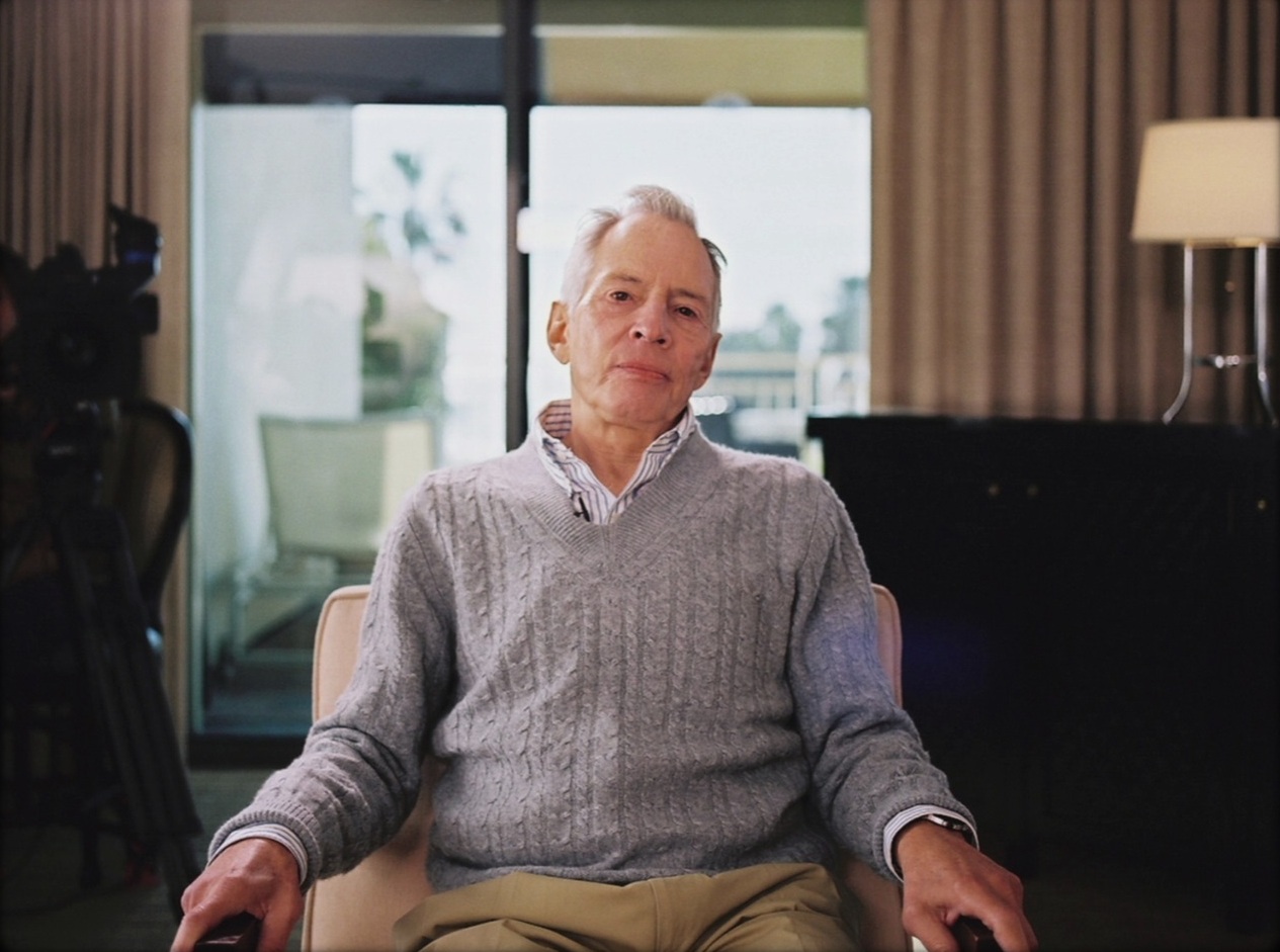 An elderly white man with white hair sits up straight in a chair, arms resting either side of him.