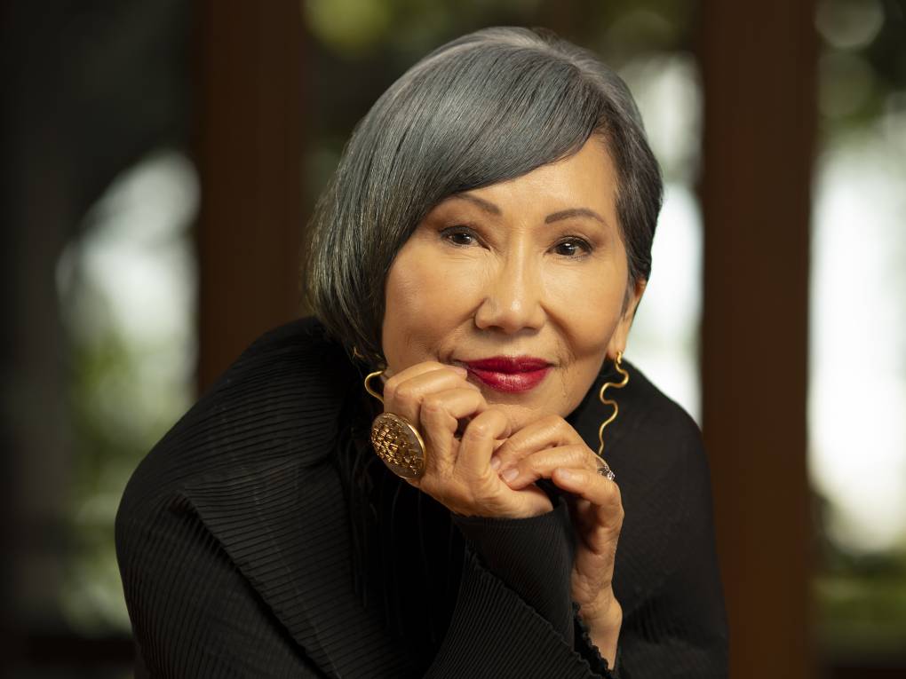 A sophisticated Asian woman with sleek grey hair rests her chin on her hands and half-smiles.